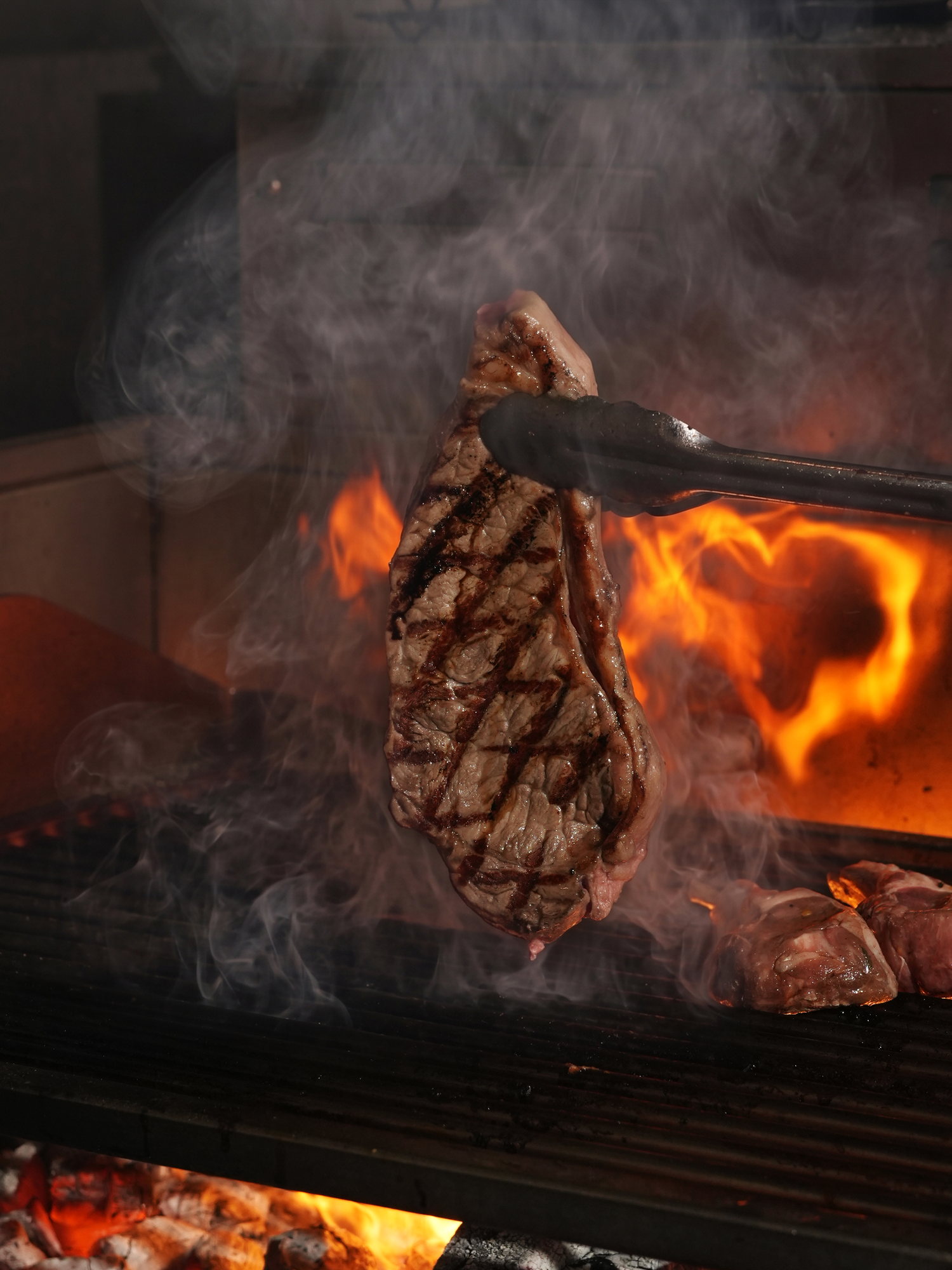 A steak with grill marks lifted with tongs over the open charcoal grill at Kanna Beach, Paradisus by Melia Bali