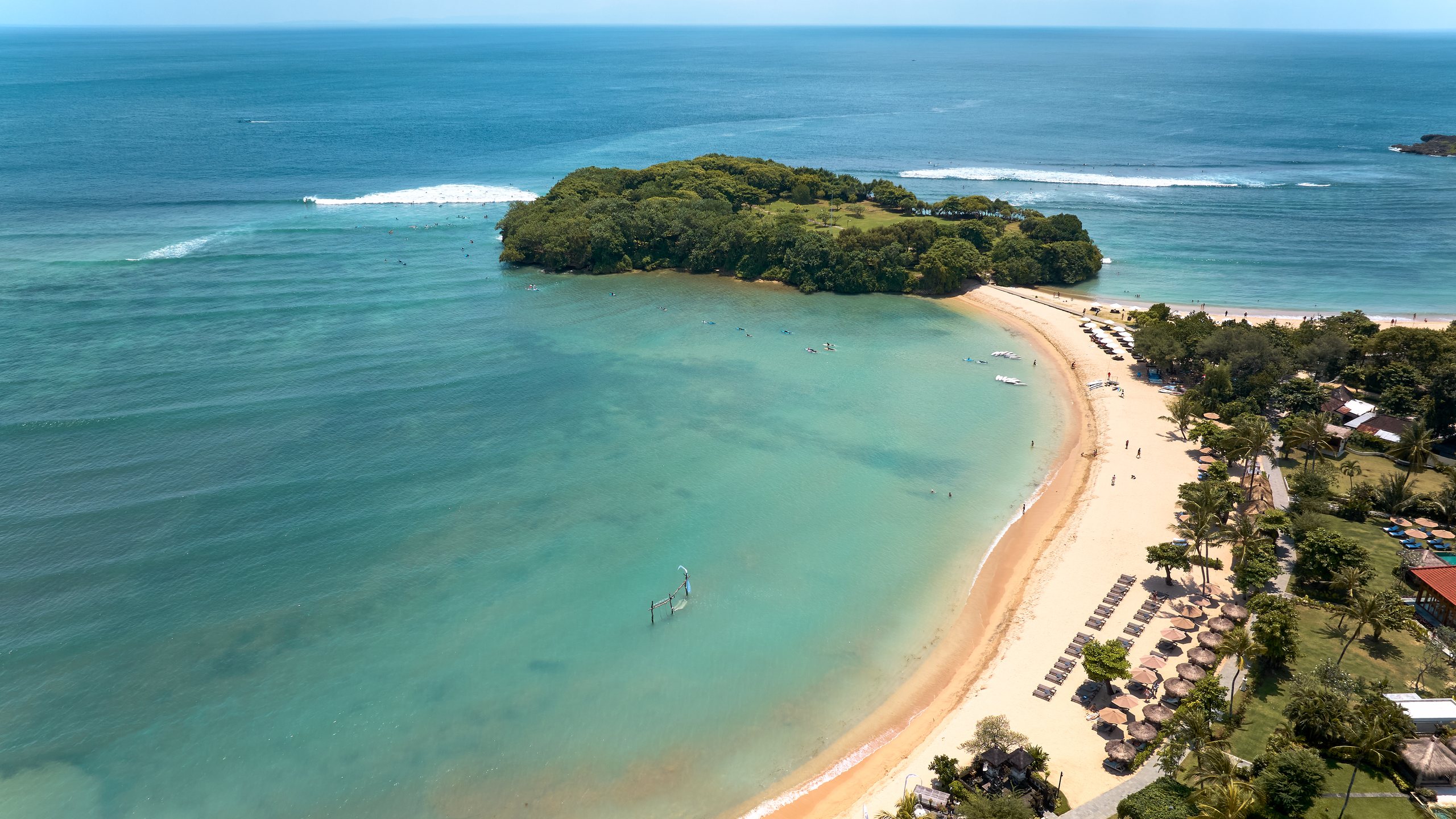 Aerial view of the beach at Paradisus by Meliá Bali in Nusa Dua: a curved sandy bay of calm turquoise water sheltered by a wooded island, with waves breaking further out