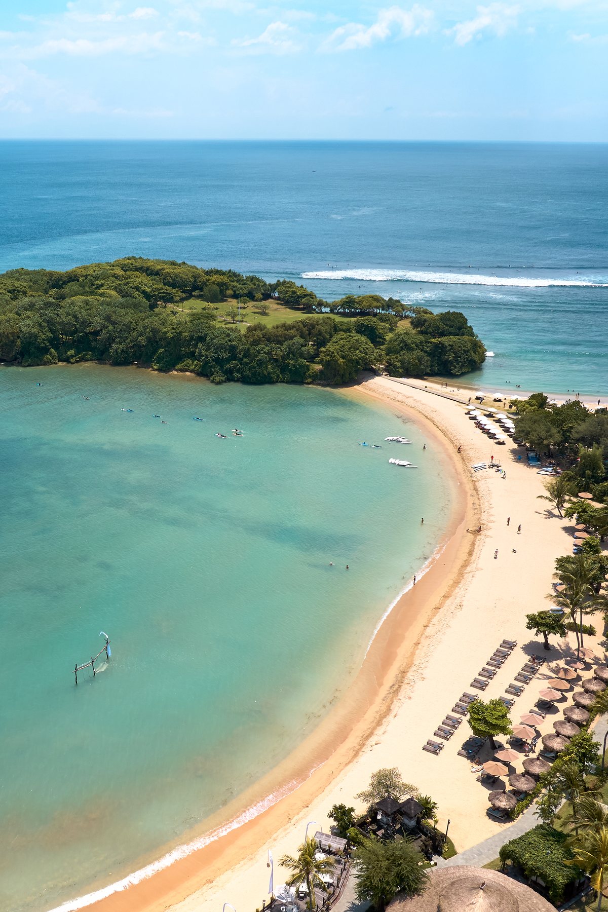 Aerial view of the beach at Paradisus by Meliá Bali in Nusa Dua: a curved sandy bay of calm turquoise water sheltered by a wooded island, with waves breaking further out