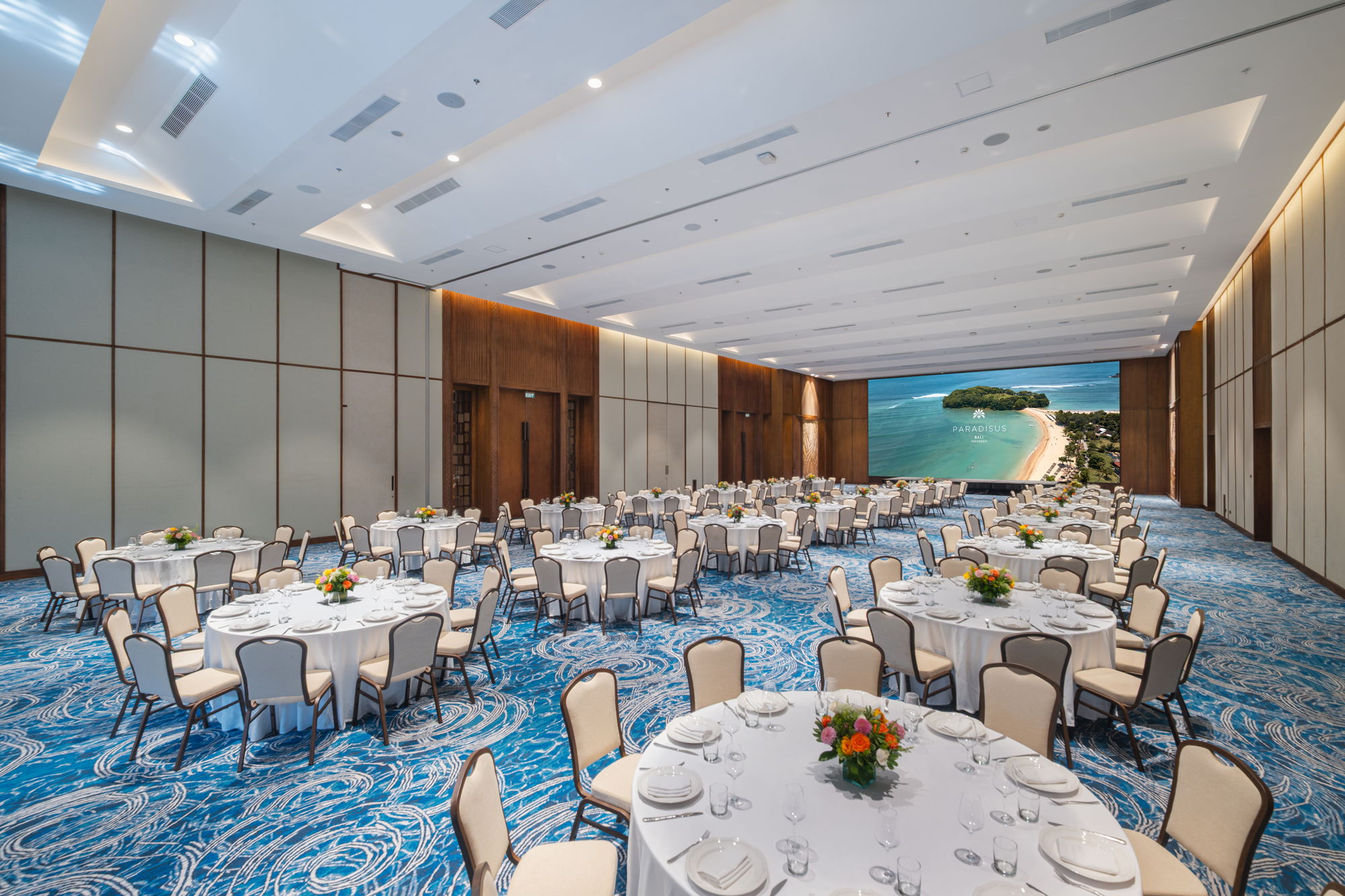 Round banquet tables set with white linen and flowers in the Puri Bali Grand Ballroom at Paradisus by Meliá Bali, with a large LED screen showing Nusa Dua beach