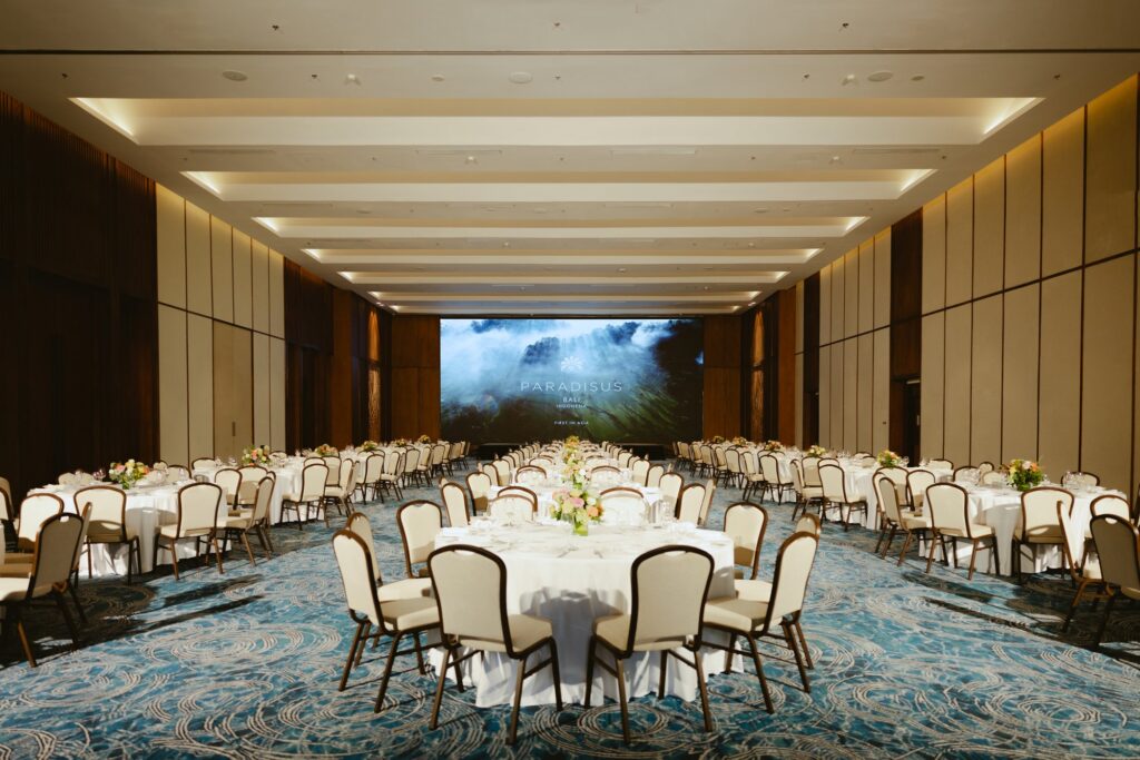 Puri Bali Grand Ballroom at Paradisus by Meliá Bali set for a banquet, with round tables in white linen, flower centrepieces and a large screen at the far end