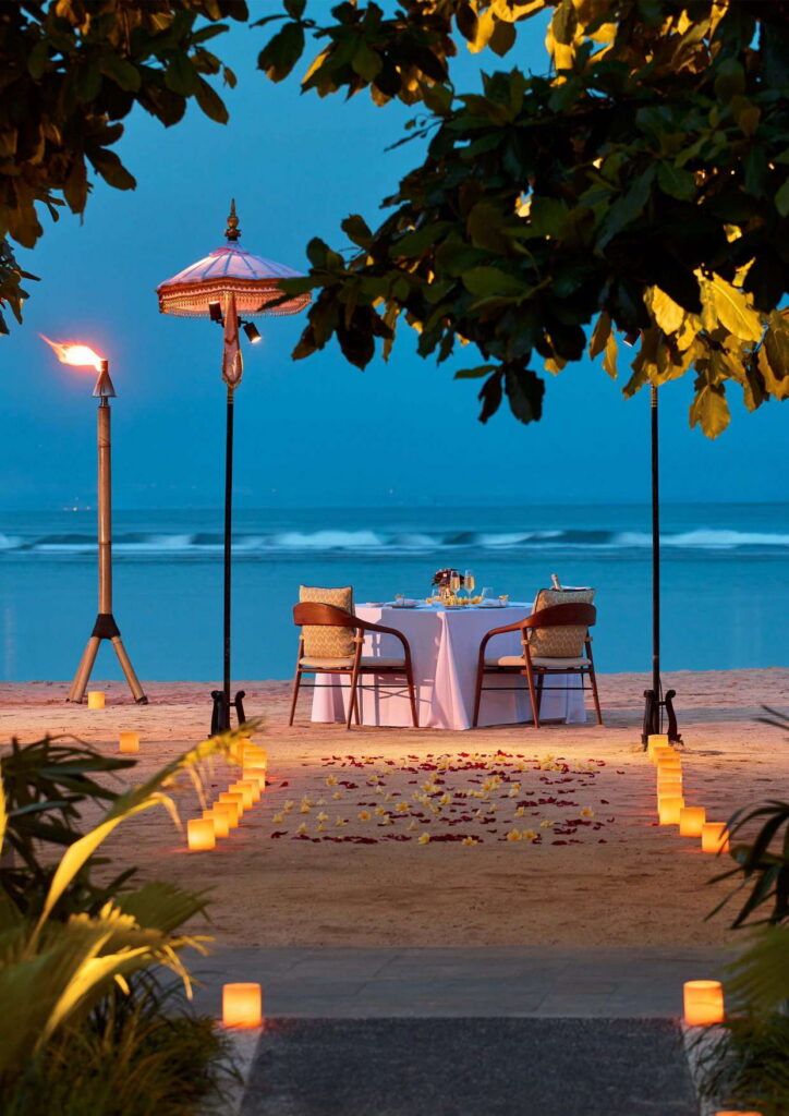 A candlelit dinner for two on the beach at Paradisus by Meliá Bali at dusk, with a Balinese umbrella and torches