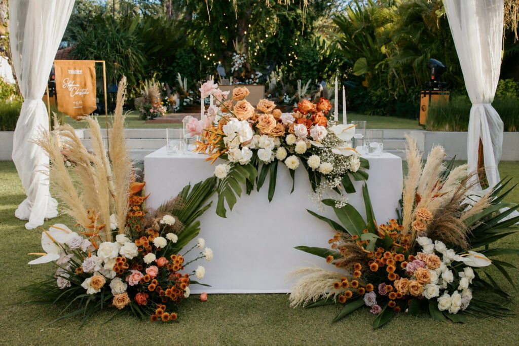 A head table in white linen on the garden lawn, dressed with peach, orange and white flowers and pampas grass between white drapes