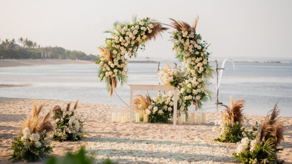 A wedding arch of white flowers and pampas grass on the beach at Paradisus by Meliá Bali in Nusa Dua, with a petal aisle, candles and the sea behind