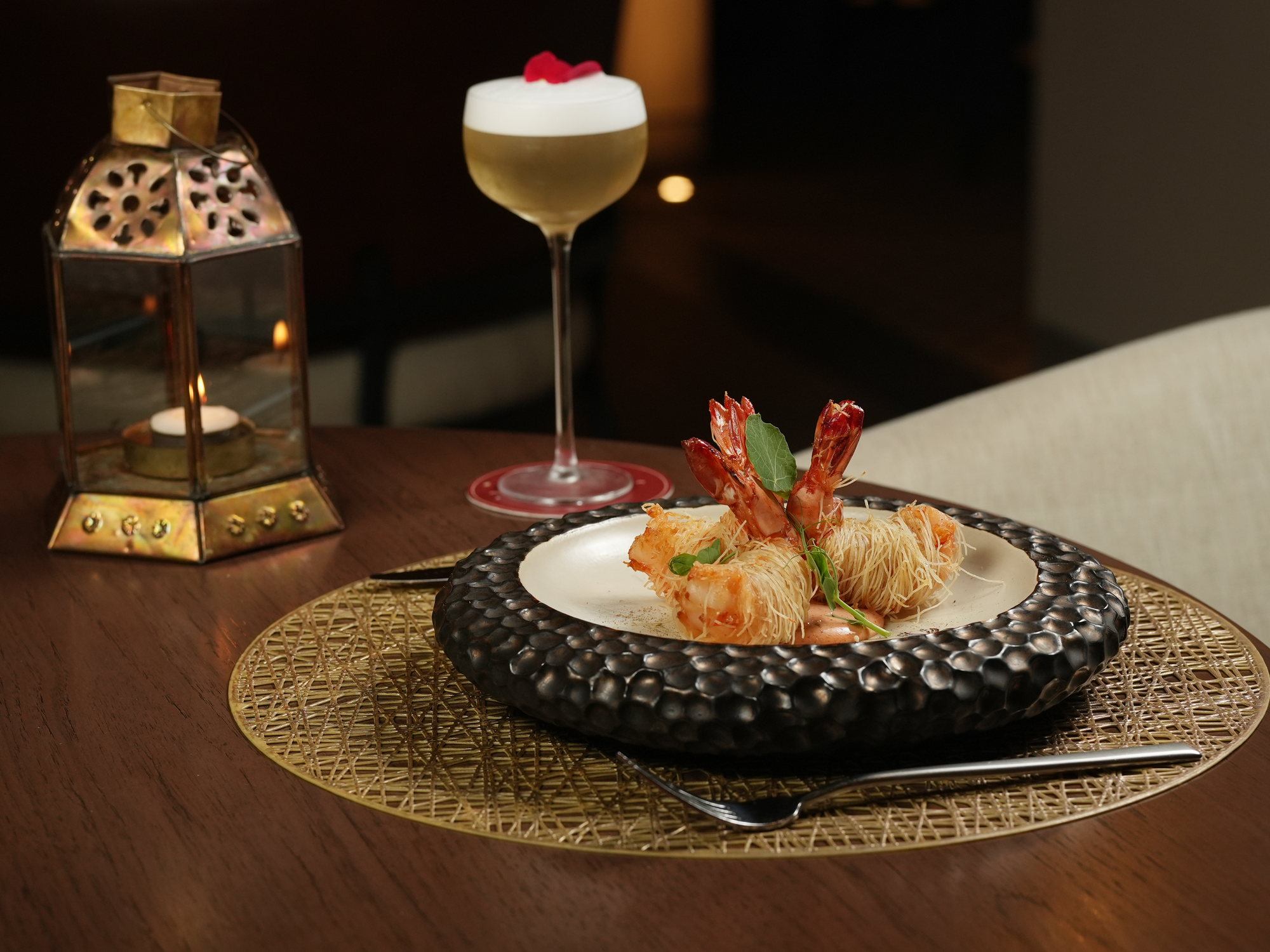Prawns wrapped in kataifi pastry on a spiced sauce, with a cocktail and a brass lantern at Samira, Paradisus by Melia Bali