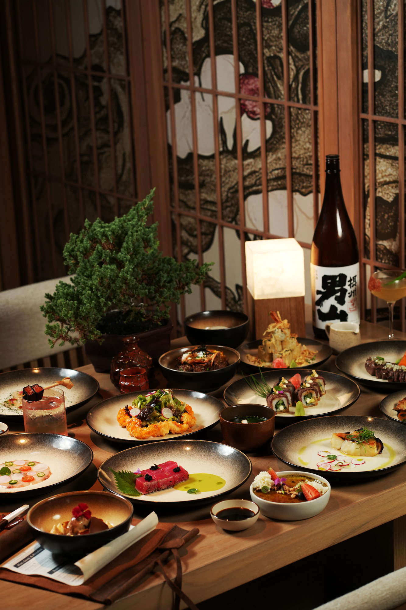 A table of Japanese dishes at Tokimeku, Paradisus by Melia Bali, laid out with a bonsai, a paper lantern and a bottle of sake against a carved screen